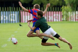 Två unga killar spelar rugby på ett gräsplan med en rugbyboll nära. Den ena har en röd och blå tröja, den andra en grön.